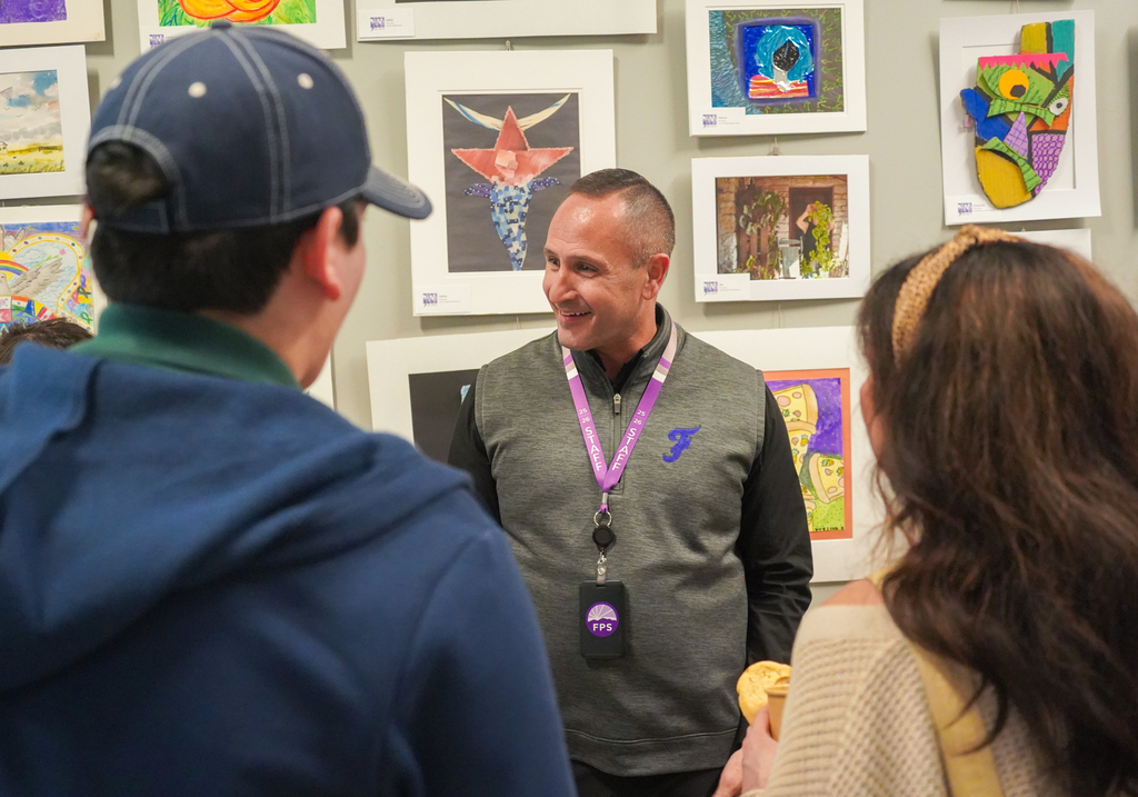 Three people stand in front of a wall with several framed artworks. One man wears a gray vest.