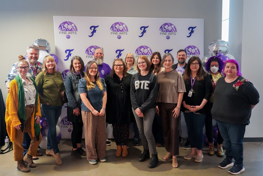 A group of adults and children standing in front of a purple and white backdrop with balloons.