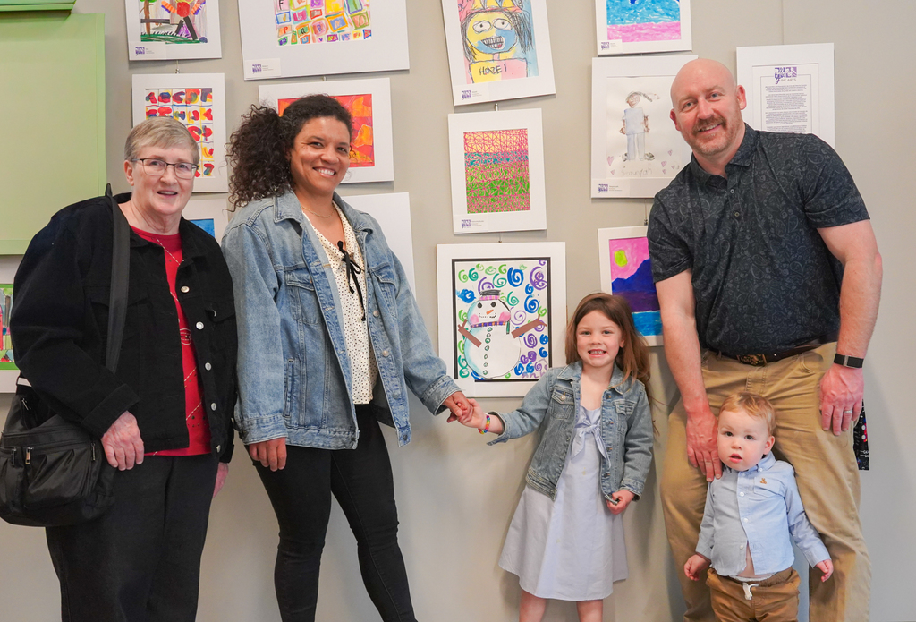 A family of four stands in front of a wall with children's artwork, smiling and holding hands.