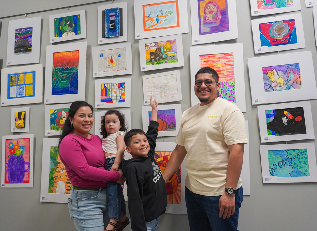 A family stands before a wall with colorful children's artwork. A woman, a child, and a boy pose together.