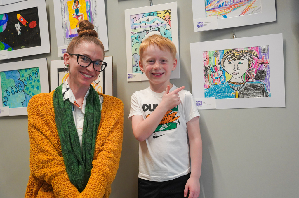 A woman and boy stand next to art pieces on a wall. The woman wears glasses and a scarf, the boy a white shirt.