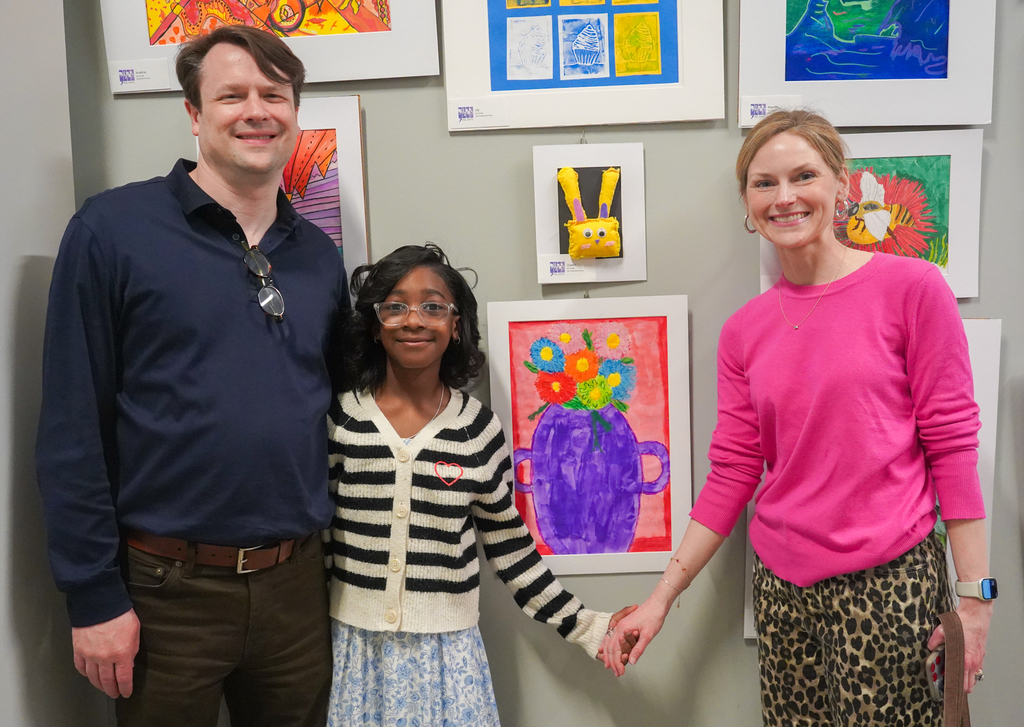 Three people stand in front of colorful paintings on a wall. A man, a woman, and a girl.