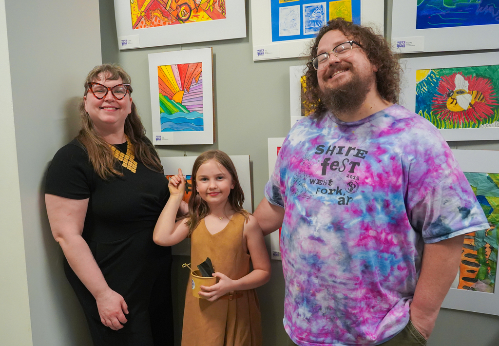 A woman and a man stand next to a girl in front of an art gallery wall with various colorful paintings.