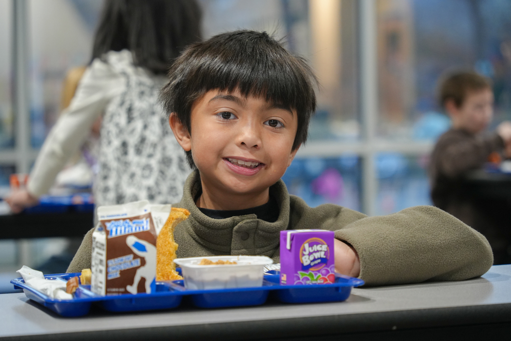 Child seated at a cafeteria table, smiling, holding a tray with food and drinks, with other children in the background.