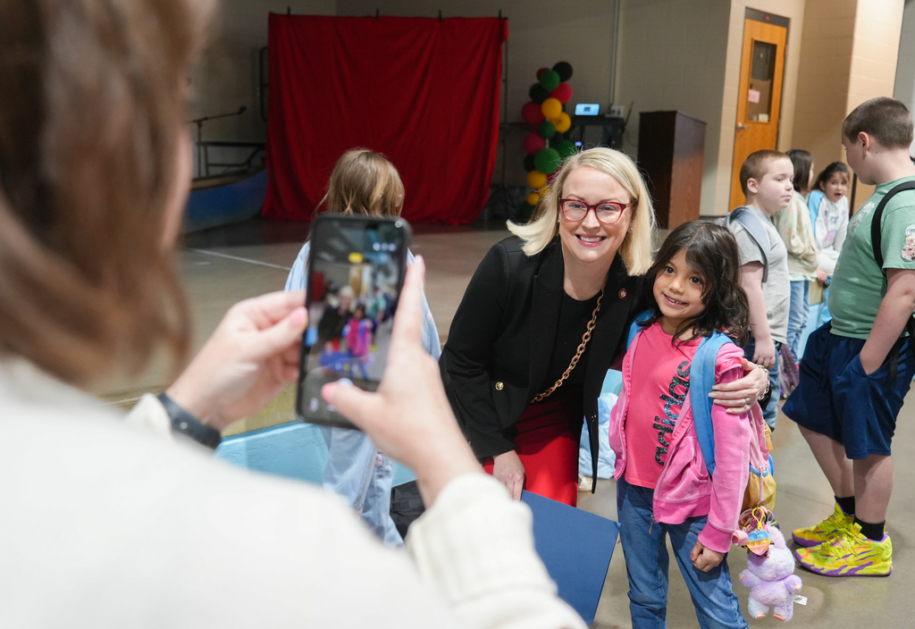 A woman holds a phone, capturing a moment of a smiling woman with a young girl. Behind them, children gather near a red curtain.