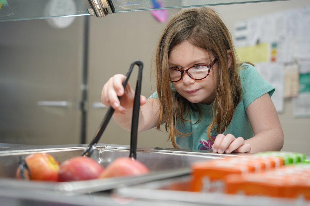 A young girl wearing glasses sits at a table, using tongs to pick apples from a buffet line.
