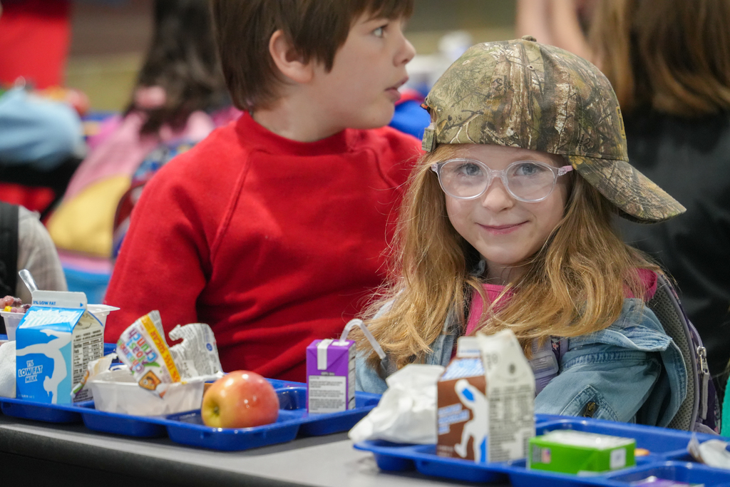 Two children sit at a cafeteria table, with one wearing a red shirt and the other a camouflage hat. Trays hold milk cartons, an apple, and other food.