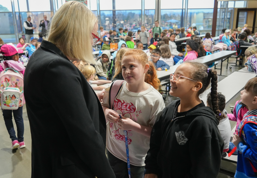 Woman speaks to two girls in cafeteria. Several children in background seated at tables.