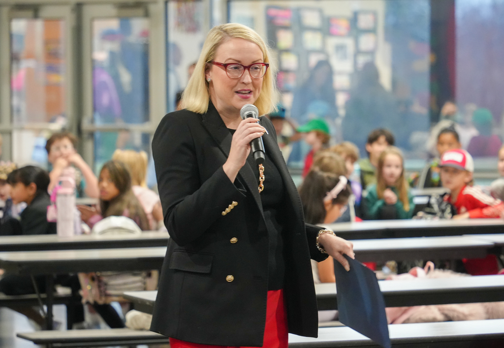 A woman with glasses speaks into a microphone in a room with students sitting at tables.