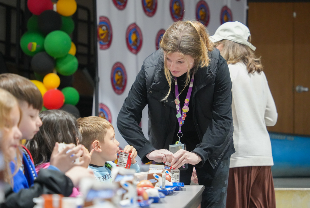 A woman stands at a table with children. She assists them with food. Colorful balloons and banners in the background.