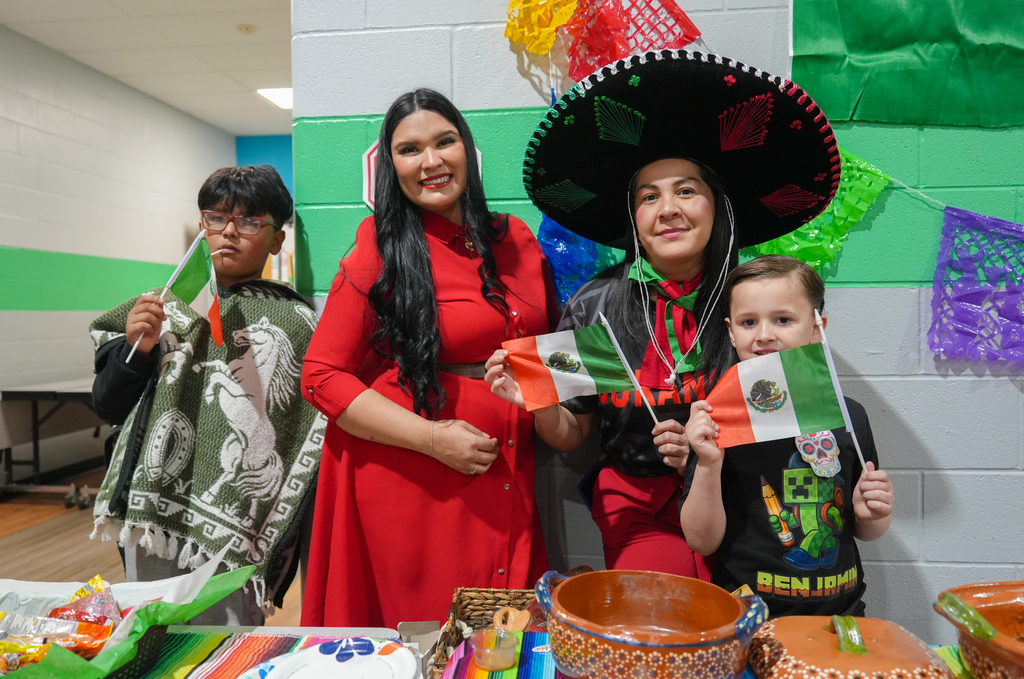 Three people stand with flags. A woman in red holds a child, and a boy in a poncho holds a flag. Behind them, a wall has colorful decorations.