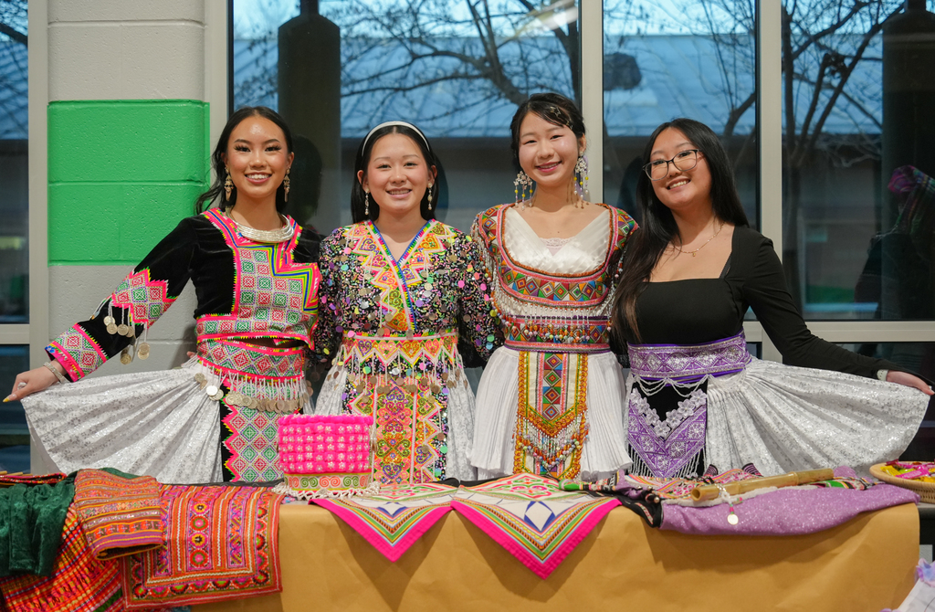 Four women in traditional attire, smiling and standing behind a table with various fabrics.