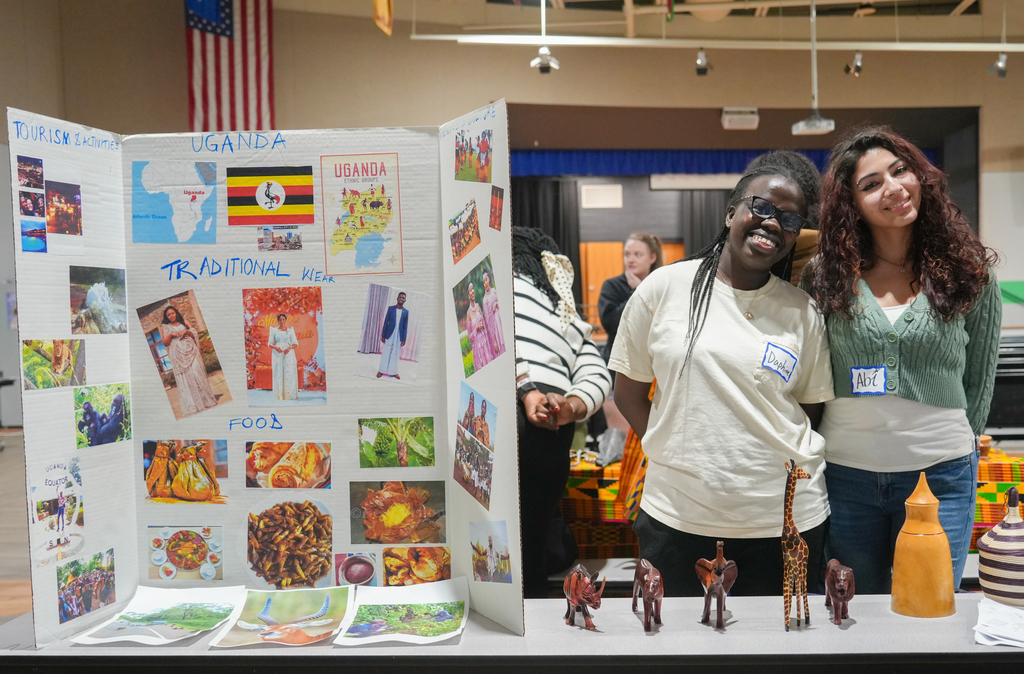 Two people stand next to a table with a display board titled "Uganda" and a flag. Various images, figurines, and papers are on the board.