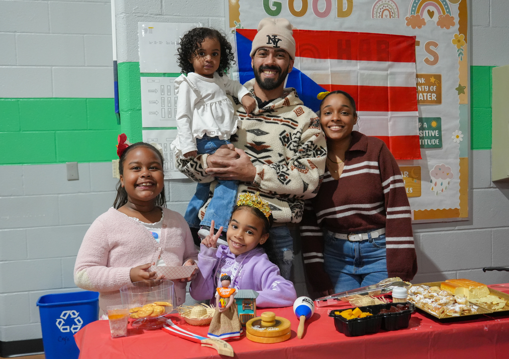 Four individuals, including a man holding a child, stand by a table with food. Two girls smile, one waves. A colorful backdrop displays text and symbols.