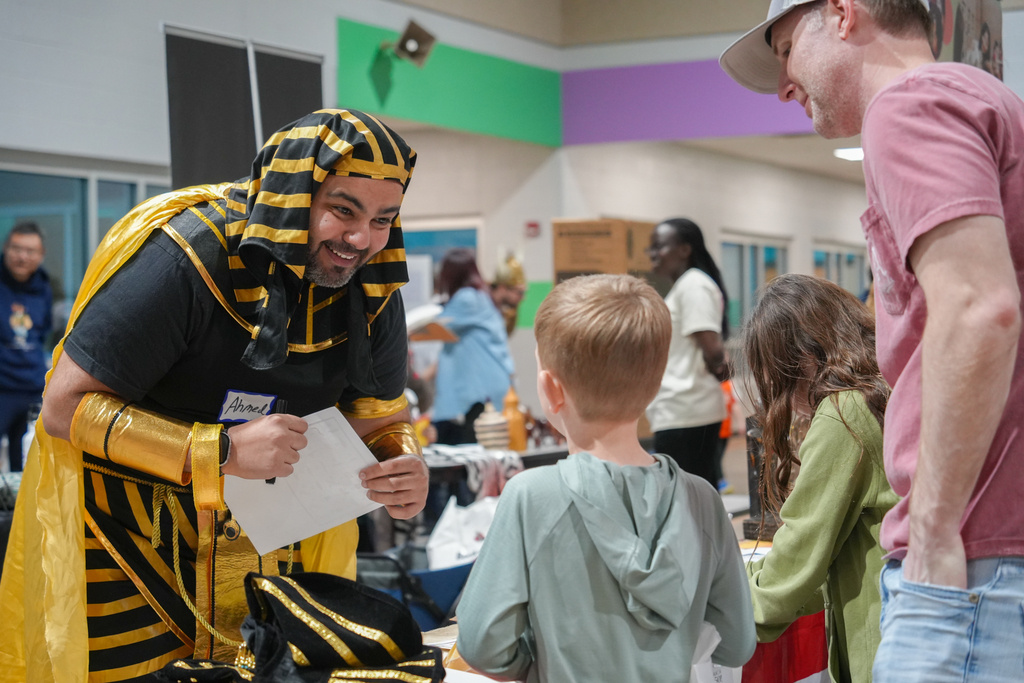 Two adults and three children, one with a backpack, engage with a person in a costume. Tables with items are visible.