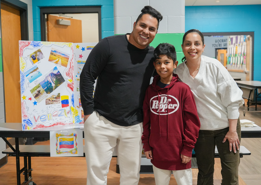 A man and two women, one in white, stand with a boy in a red hoodie. Behind them, a table holds colorful artwork.