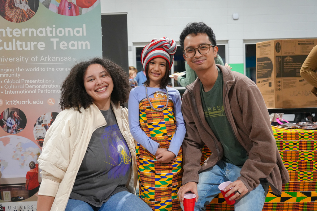 Three people sit on a bench with a colorful cloth backdrop. A smiling woman, a child, and a man.