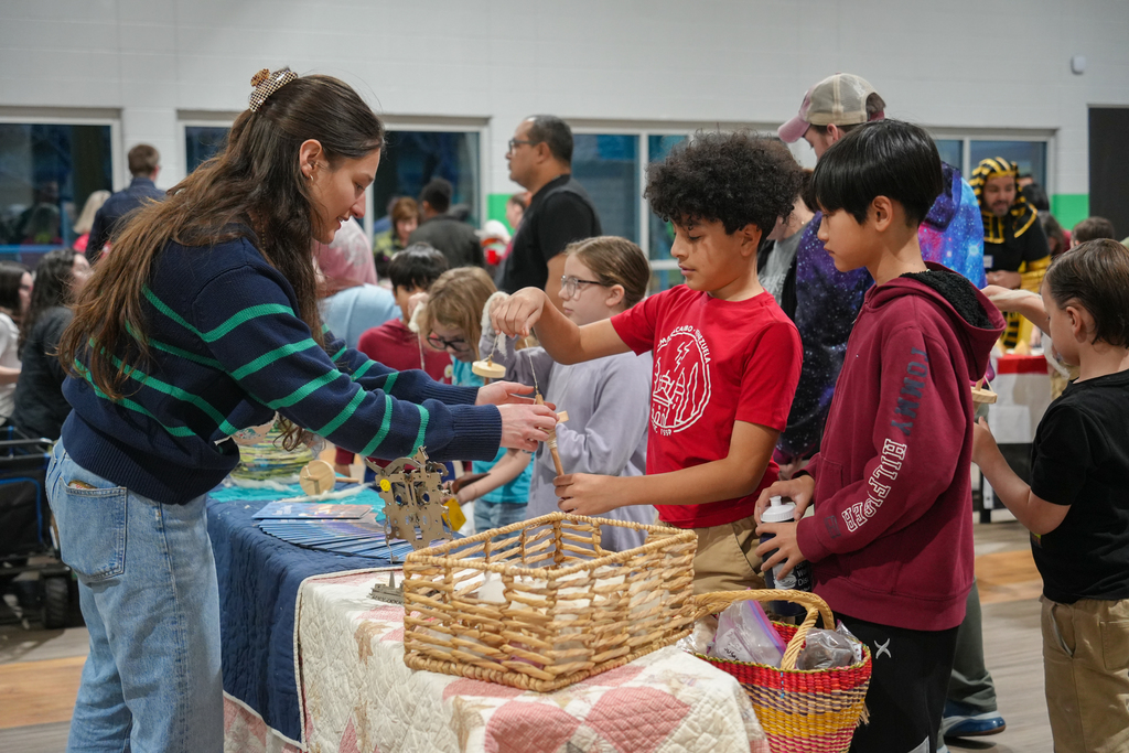 A woman assists children at a table with baskets, others are gathered around. The room has windows and a plain wall.