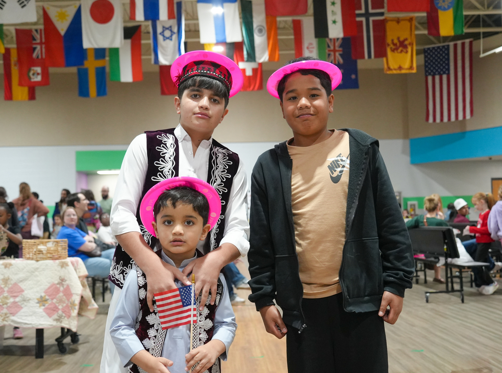 Three children wear pink hats; one holds a flag. Behind them, a large room with flags, tables, and people.