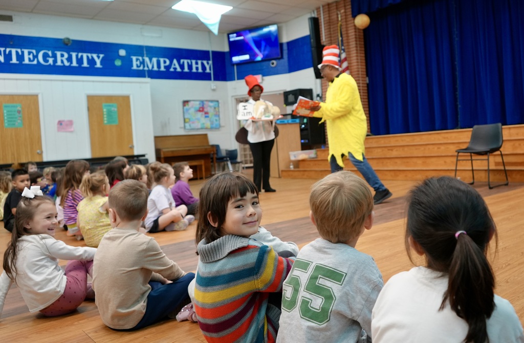 Children sit in a classroom with a stage, a person dressed in a yellow costume performs, and a piano is nearby.