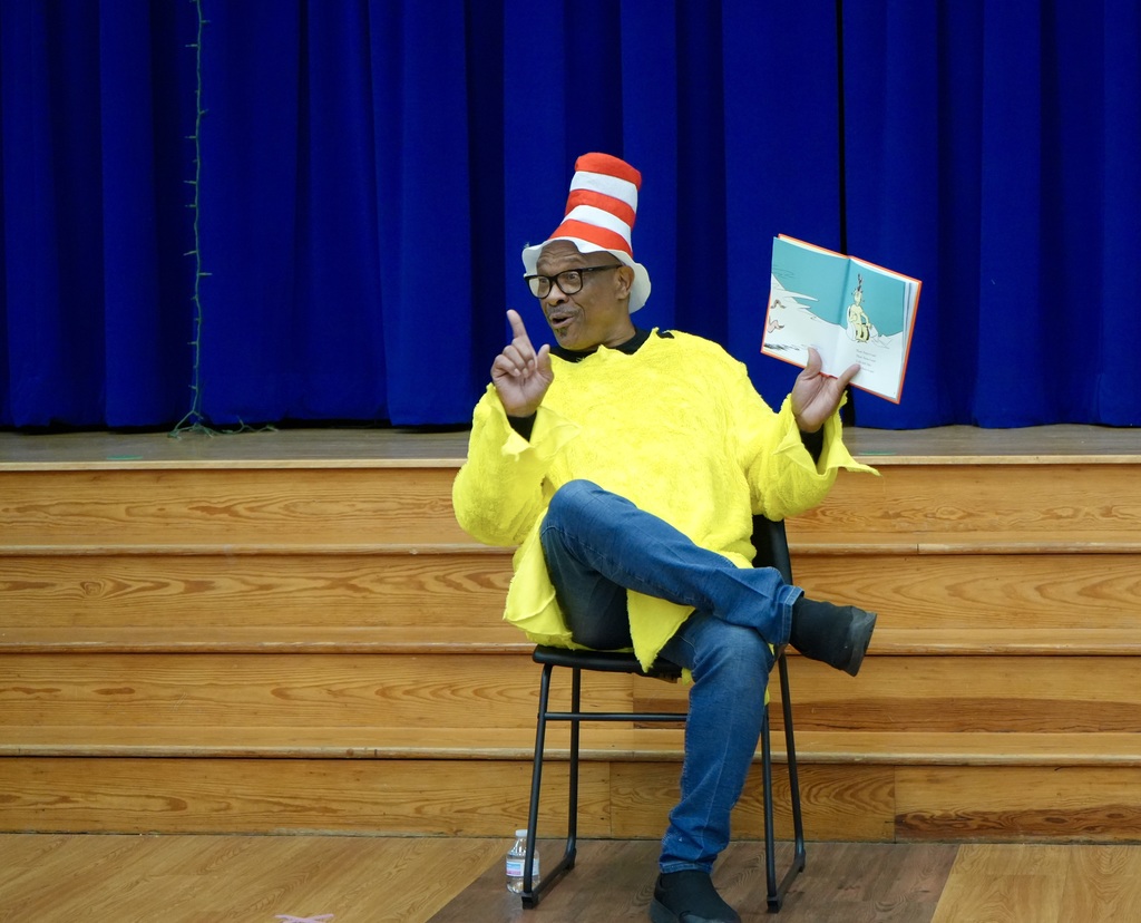 A man in a yellow Cat in the Hat costume is sitting on a chair, holding a book.