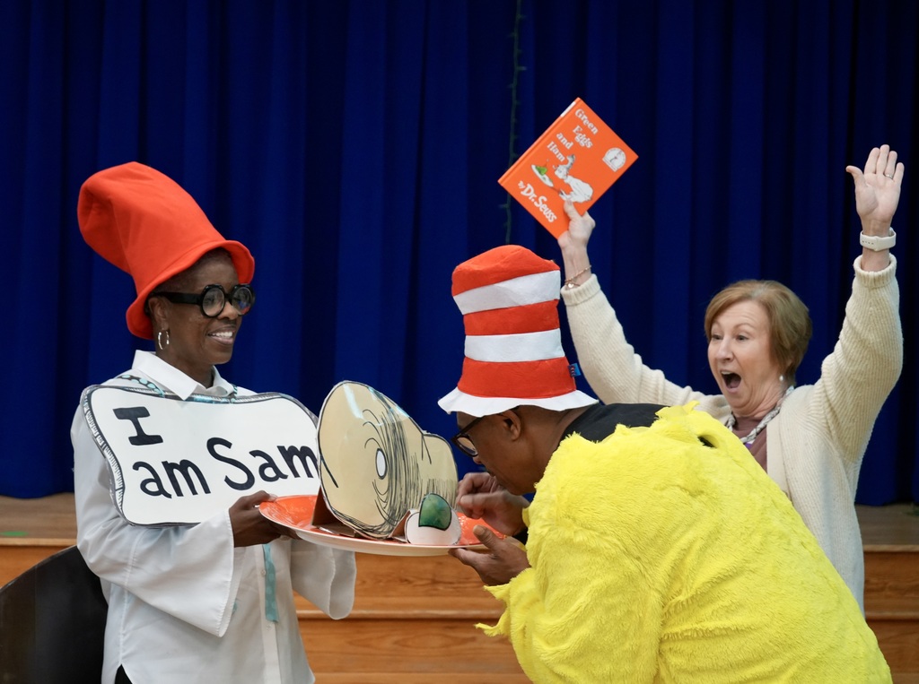 Three people dressed in Dr. Seuss-inspired costumes on a stage, holding signs and waving. Background includes a blue curtain.