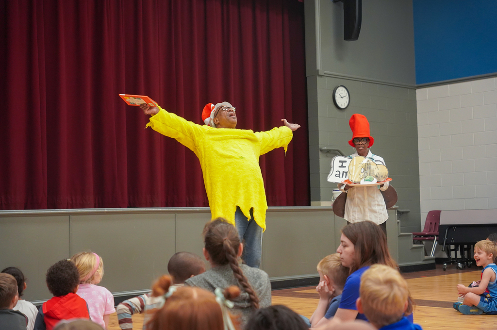 Two adults perform a play for a group of children. The adults wear hats and costumes. The children watch.
