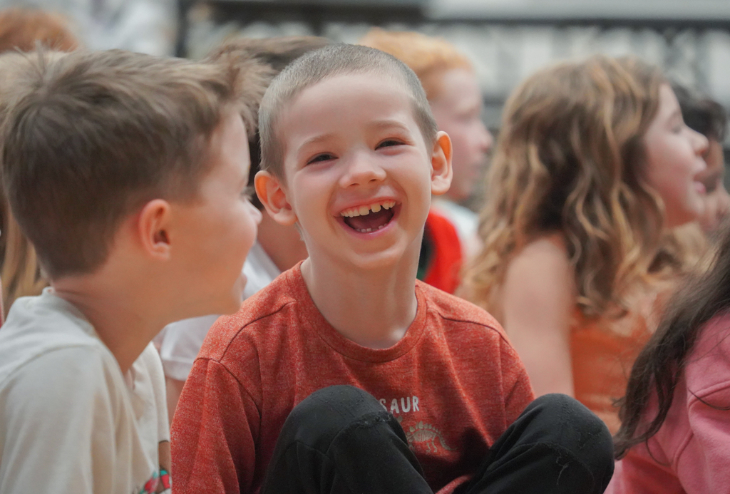 Kids in a group, one boy in red with a big smile looking at another boy in white.