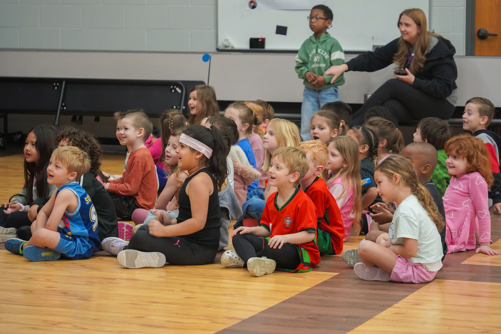 Group of children seated on a gymnasium floor, listening to a woman. A whiteboard is visible in the background.