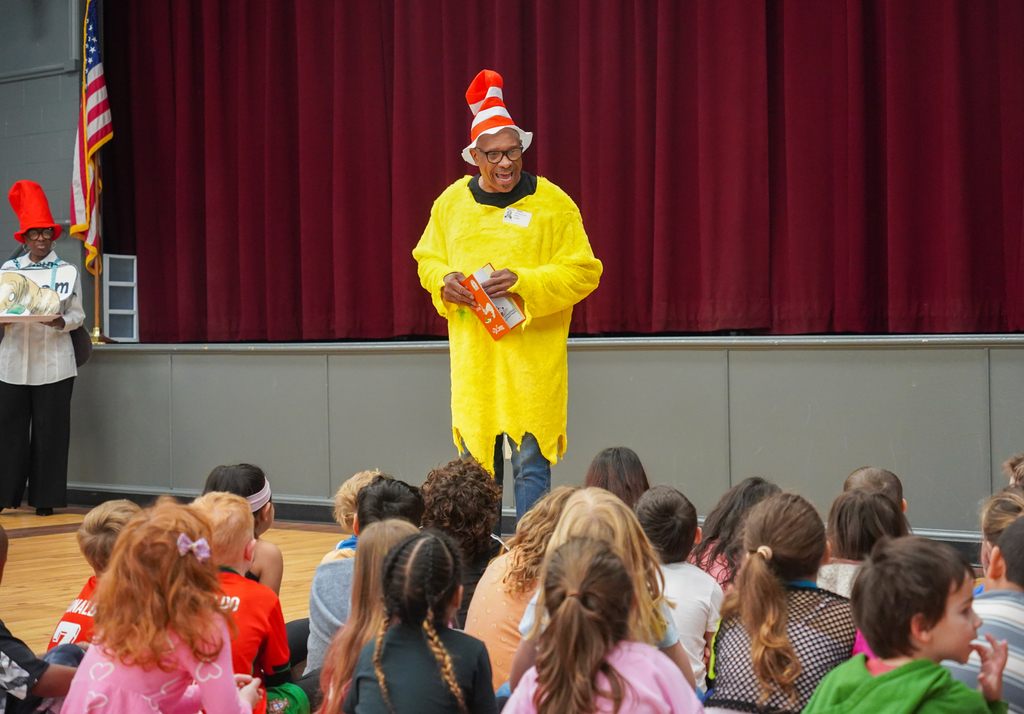 A person in a yellow costume and a red hat reads to a group of children.