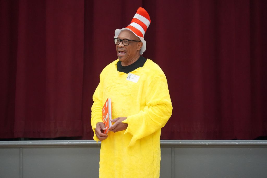 Person in yellow garb and red-white striped hat, holding a book, stands on a stage with red curtains.