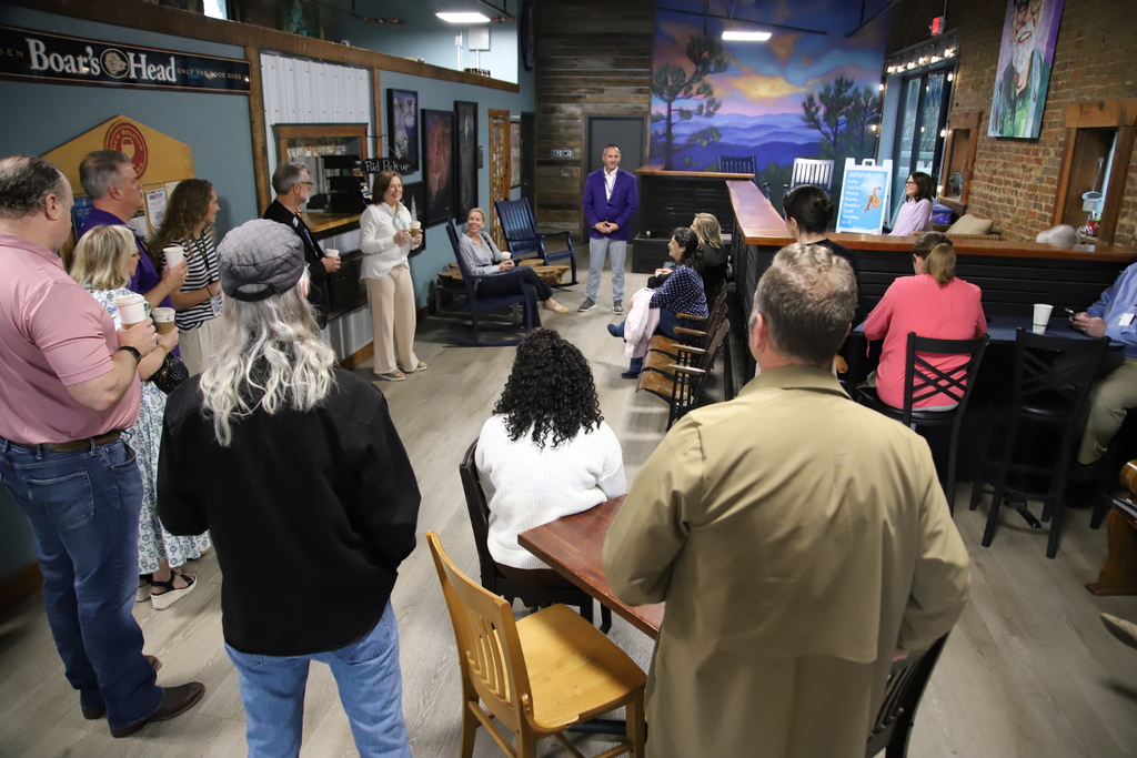A group of people in a room with a stage, wooden floor, and tables. Some sit, others stand.
