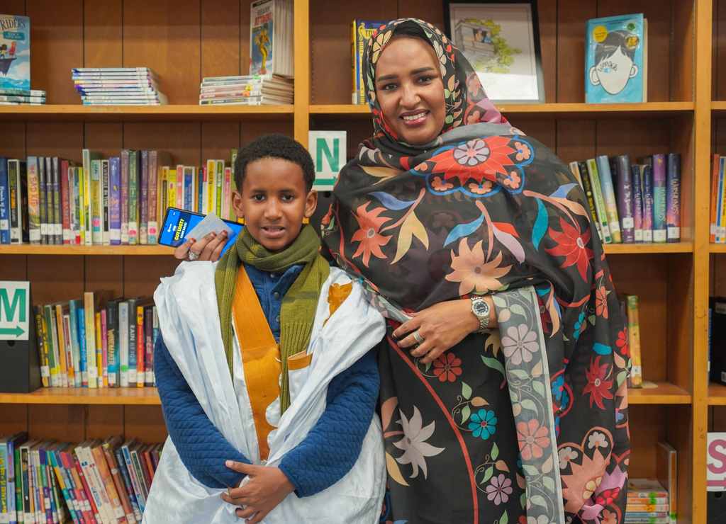 Woman in patterned dress and boy in white garment standing in front of bookshelves, woman with hand on boy's shoulder.