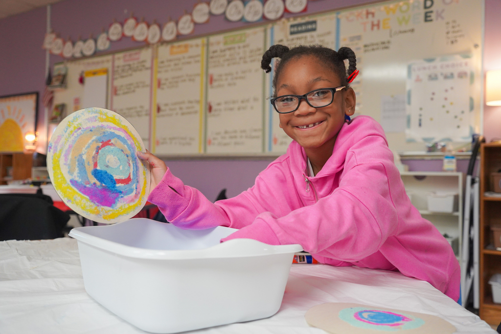 Child holds artwork at table with tray. Bright classroom background with whiteboards.