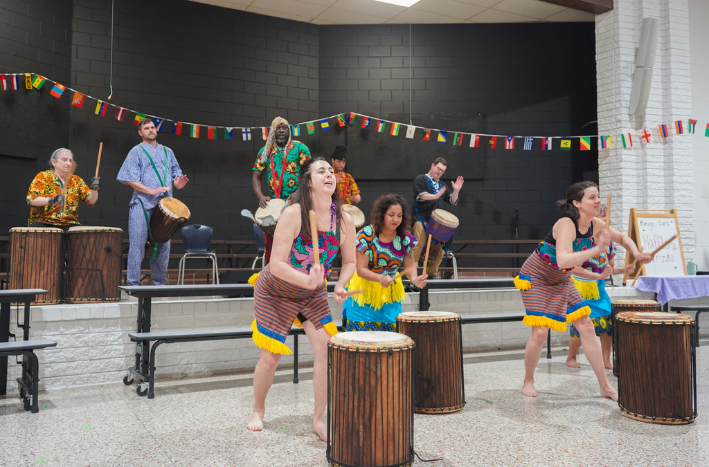 A group of people play drums in a room with multicolored flags and a table with objects.
