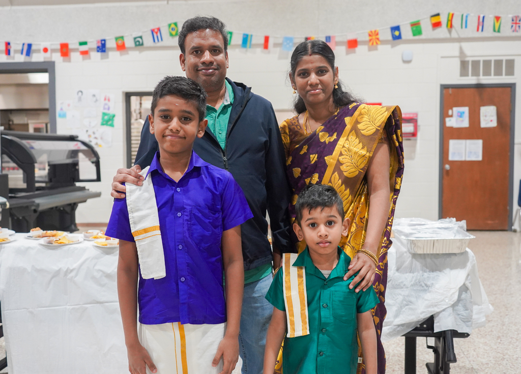 Four individuals in traditional attire stand together; a man and two boys, with a woman beside them. Behind them, tables with food and flags adorn the space.