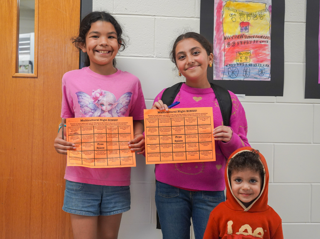 Two girls in pink shirts and a boy in a red hoodie pose with orange papers. One girl wears a backpack.