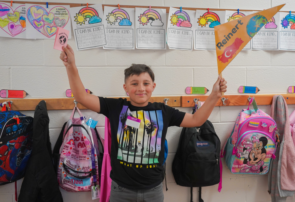 A boy with a smile holds a flag and a card. He stands among colorful backpacks and jackets on a wall.