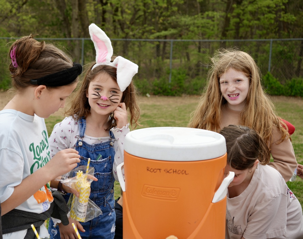 Four young girls, some with face paint, gather around an orange container. One holds a drink, and a blurry fence and trees are in the background.