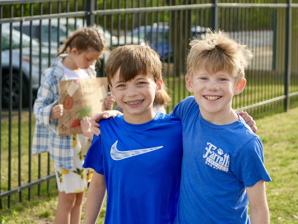 Two boys smile and stand next to each other, wearing matching blue shirts. A girl stands behind them, holding a paper bag. Behind them, a black fence and a parked car.