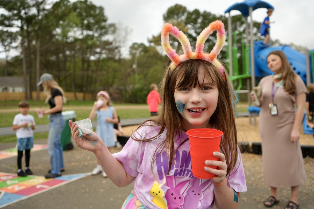 A girl with bunny ears holds a red cup in a park with children and adults.