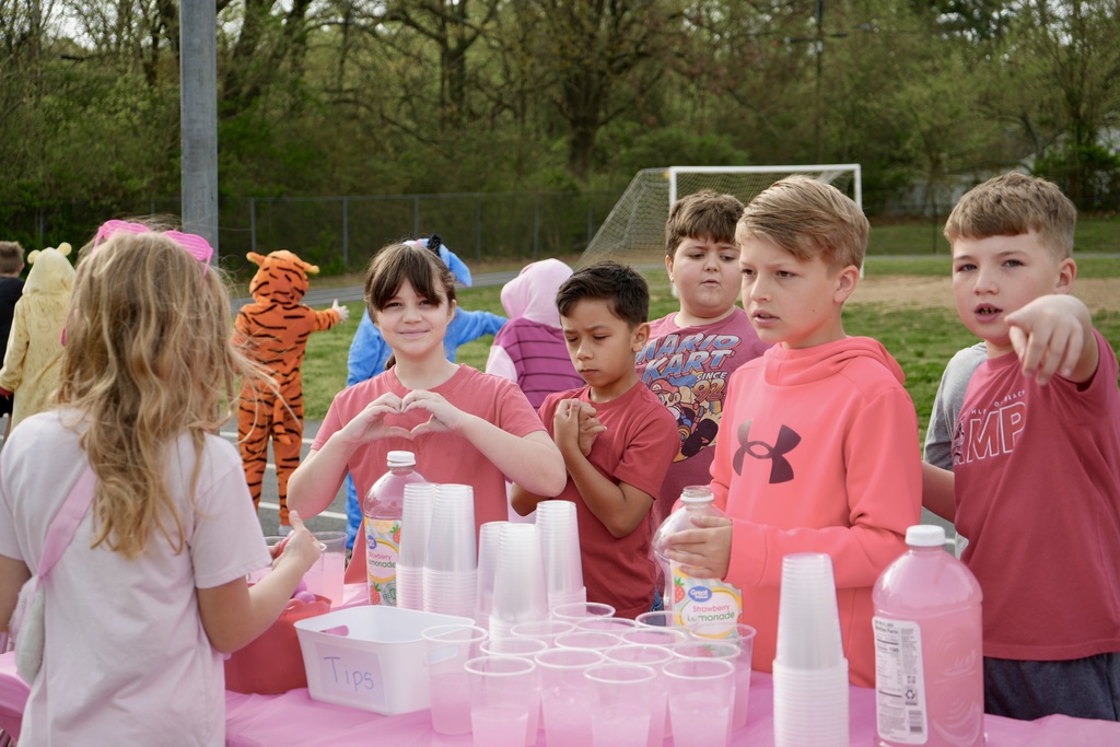Kids dressed in pink stand at a table with cups and bottles; a soccer goal is in the background.