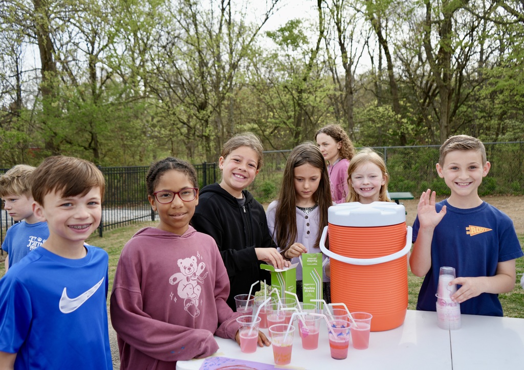 Group of children stand behind a table with cups of red drink. Trees in background.
