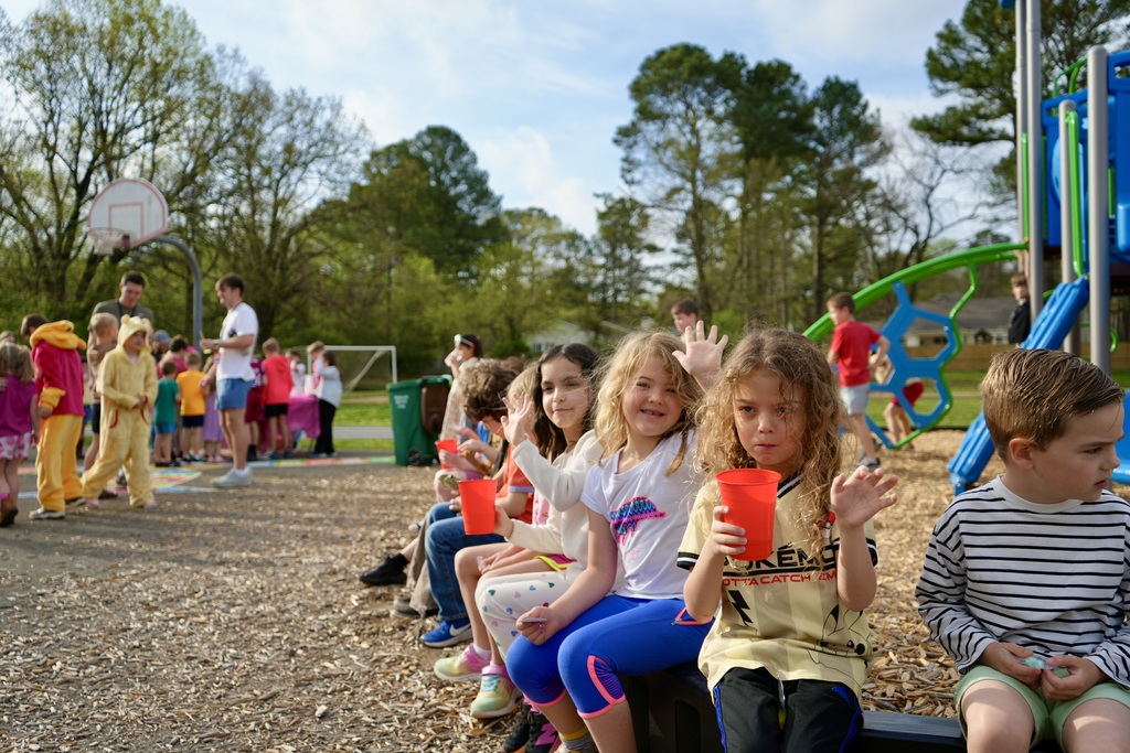 Children sit in front of a playground with trees and a basketball hoop in the background.