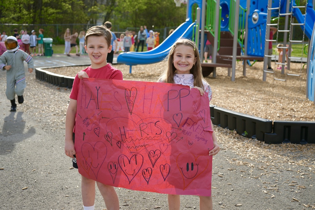 Two children hold a heart-filled sign in a playground with a slide and other play equipment.