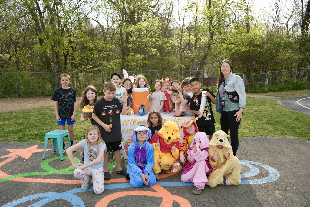 Kids in costumes and an adult stand in a circle on a playground. A table with items is behind them.