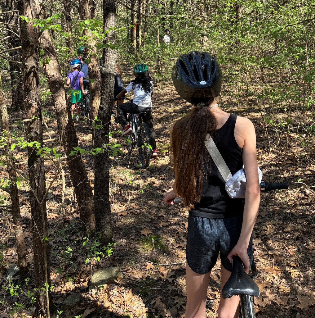 A woman in a forest biking trail, with others wearing helmets and biking behind her.