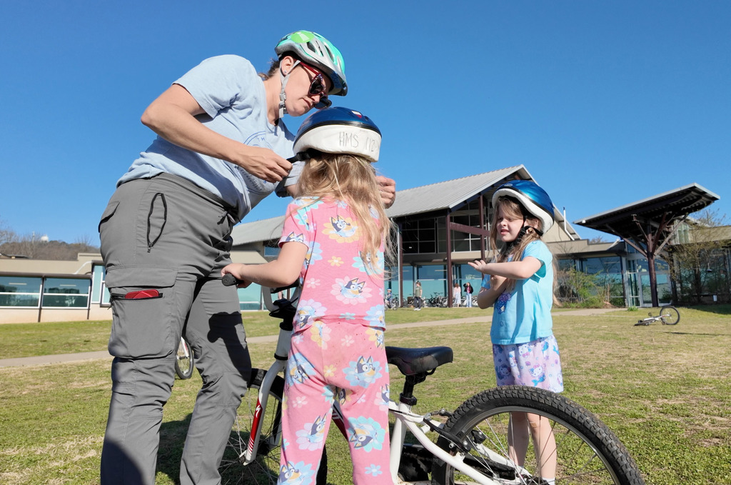 An adult helps two children put on helmets. They stand on grass with a building in the background.