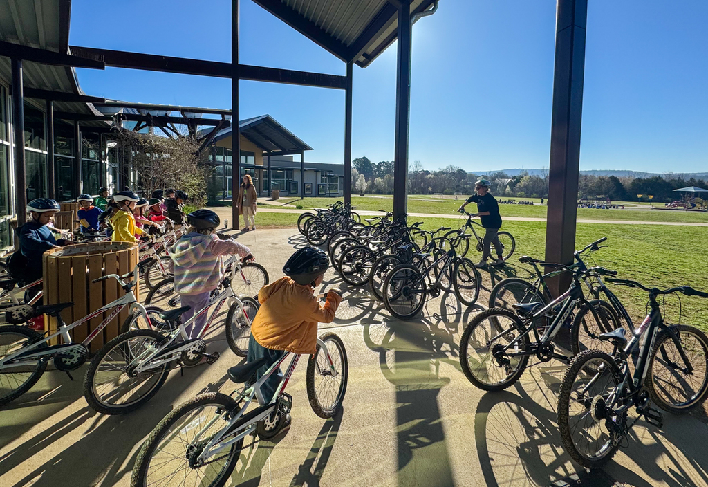 Children in helmets ride bicycles in a line under a covered area with a grassy field in the background.