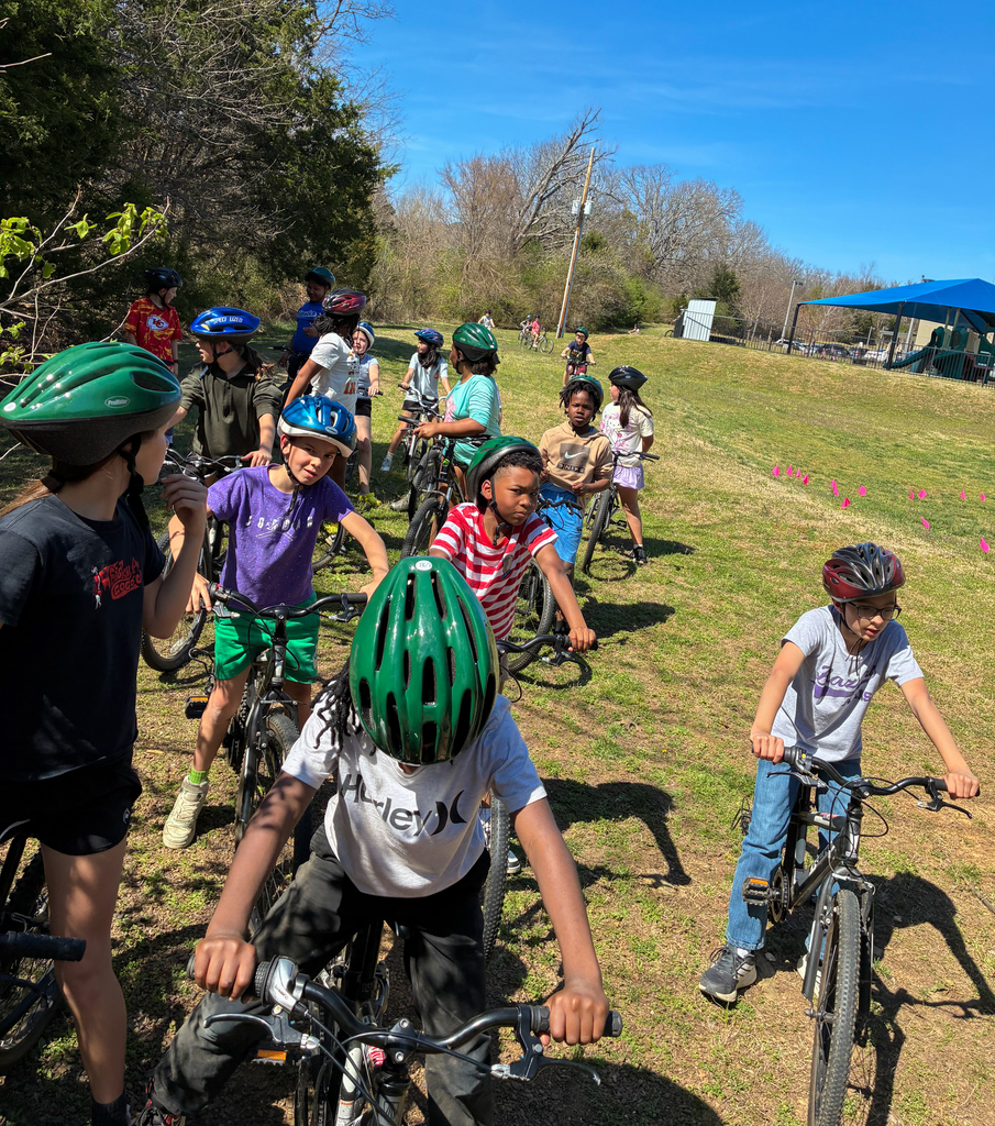 Children in helmets on bicycles in a grassy area. A few children wear matching colors.